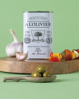 Olivenöl in Blechdose von A L'Olivier auf Holzbrett mit Knoblauch und Oliven, mediterrane Atmosphäre vor grünem Hintergrund