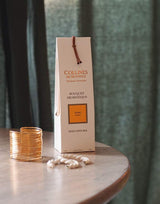 Collines de Provence Duftdiffuser Amber Verpackung auf einem runden Tisch neben einem geriffelten Glas und einer weißen Kette