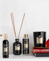 Collines de Provence Duftkollektion mit Raumspray Ebenholz, Duftdiffuser Ebenholz, Duftdiffuser-Nachfüllung Schwarze Rose und Duftkerze Zeder auf Regal mit Büchern und roter Blüte vor hellem Hintergrund.