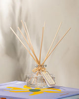 Durance Raumduft Diffuser Pudrige Veilchen mit dekorativem Glasflakon und Duftstäbchen auf lilafarbenem Buch in stilvoller Wohnraumdekoration mit Duftbezeichnung Violette Poudrée Powdered Violet