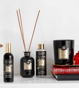 Collines de Provence Duftkollektion mit Raumspray Ebenholz, Duftdiffuser Ebenholz, Duftdiffuser-Nachfüllung Schwarze Rose und Duftkerze Zeder auf Regal mit Büchern und roter Blüte vor hellem Hintergrund.