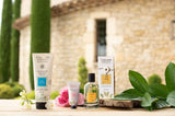 Collines de Provence Pflegeserie mit Duschgel Tiaréblüte, Handcreme Rose von Grasse sowie Eau de Toilette Amber, dekorativ arrangiert vor mediterraner Kulisse.