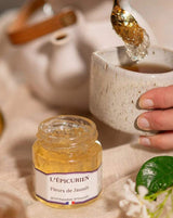 Serviervorschlag mit L'Épicurien Jasminblüten-Gelee: Ein geöffnetes Glas steht neben einer Tasse, während ein Löffel mit Jasminblüten-Gelee darüber gehalten wird.